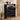 Black shoe cabinet with shoes on a wooden floor, next to a plant and a door.