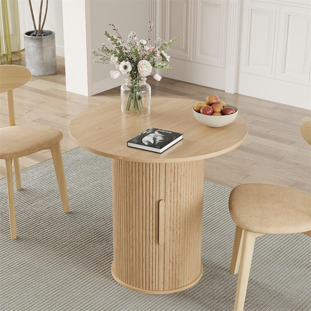 Round wooden table with a vase of flowers and a bowl of fruit in a room with chairs and a plant.
