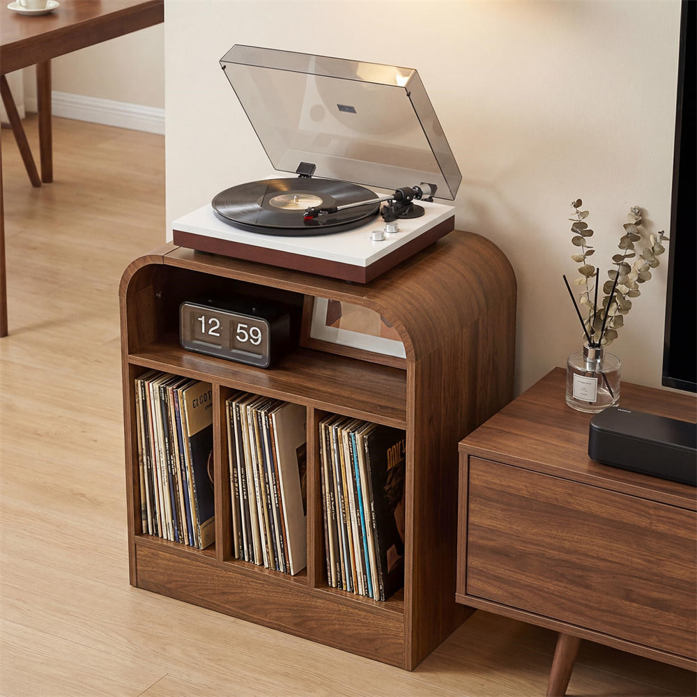 A mid-century modern wooden record player console with a turntable on top, open shelves holding vinyl records, and a digital clock, shown in a living room setting next to a matching wooden side table.