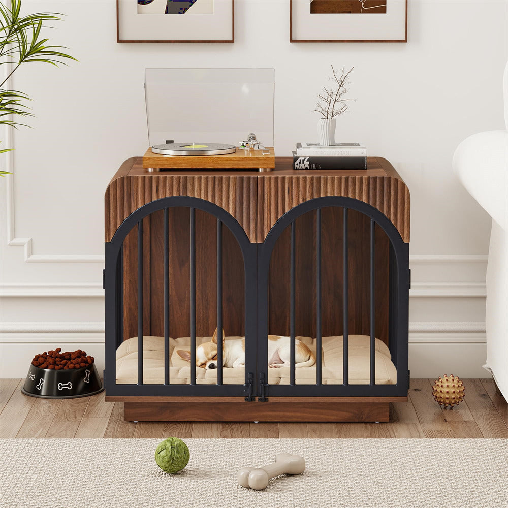 A wooden pet crate with black metal double doors and a cushioned bed inside, shown in a styled home setting with a dog resting underneath, a turntable on top, and pet toys on the floor.