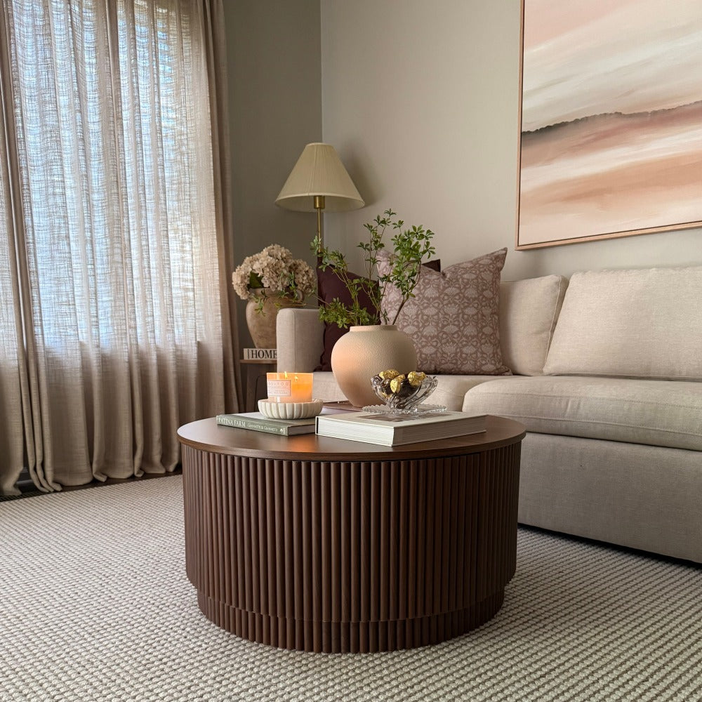 A round coffee table with a fluted dark brown wood finish sits in a modern living room setting next to a light beige sofa and decorated with vases and candles.