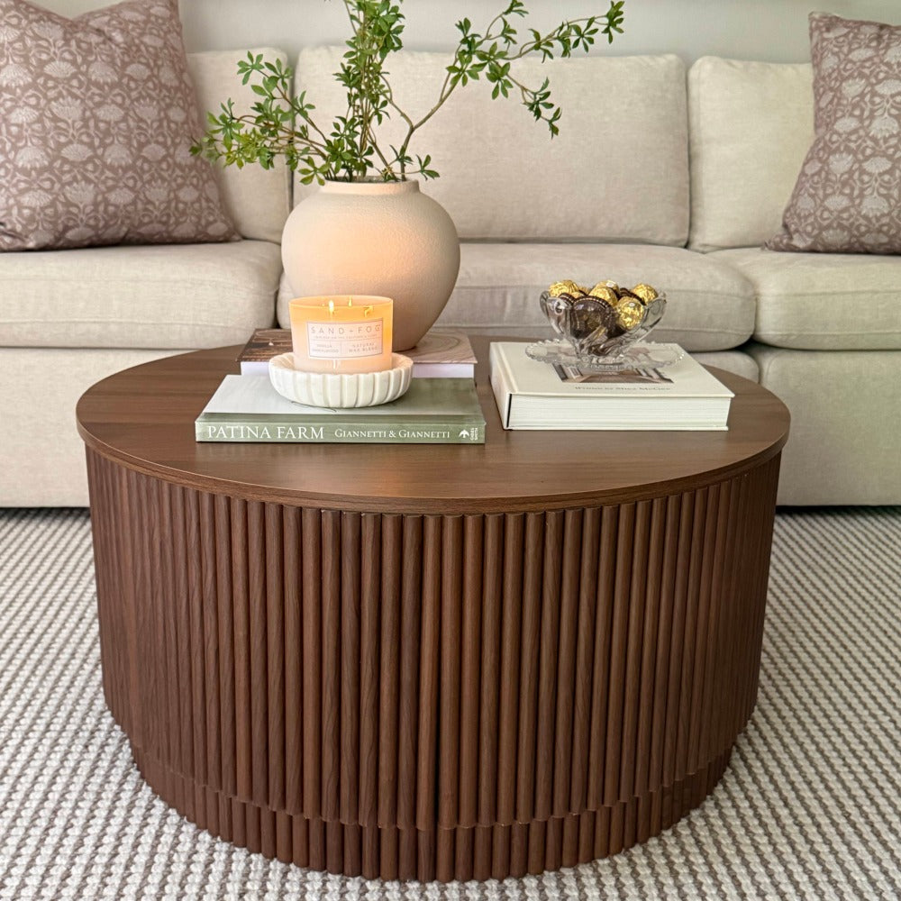 A round wooden coffee table with a fluted slatted front, styled in a living room setting with a beige sofa, patterned pillows, a ceramic vase with greenery, books, and candles on top.