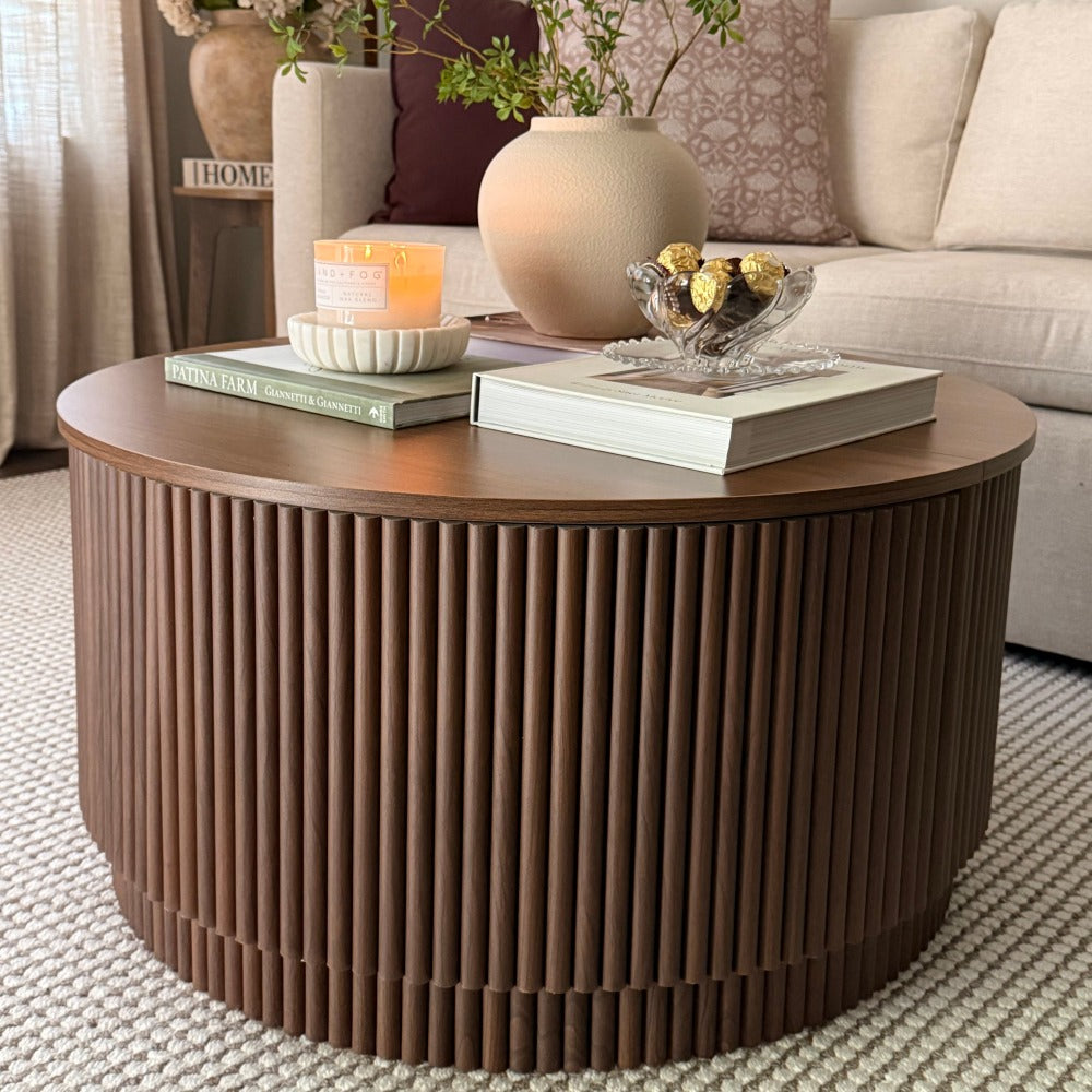 A round coffee table with a fluted vertical slat design and a medium brown wood top, styled in a living room setting with books, a candle, and a vase.