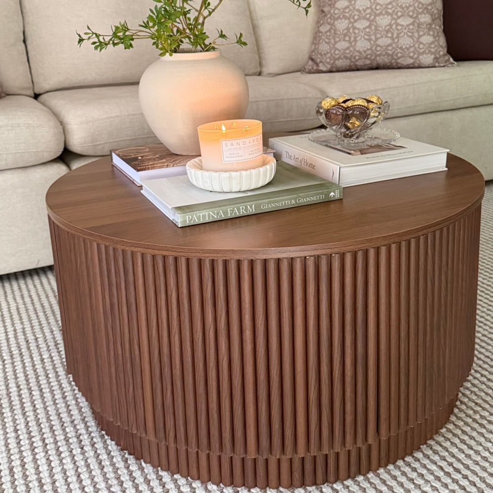 A round wooden coffee table with a fluted slatted front, styled in a living room setting with books, a candle, a vase, and a glass bowl on top.