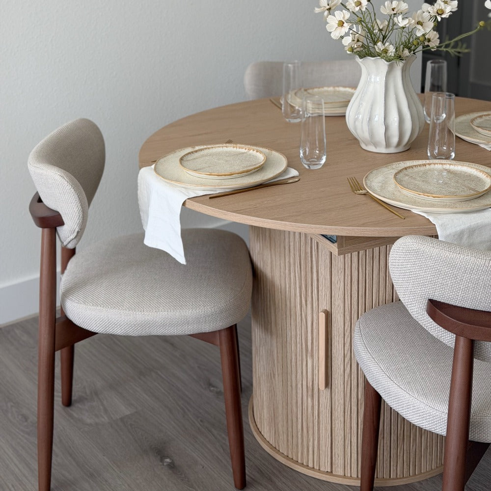 A round light wood dining table with a fluted pedestal base, surrounded by upholstered dining chairs in a bright dining room setting.
