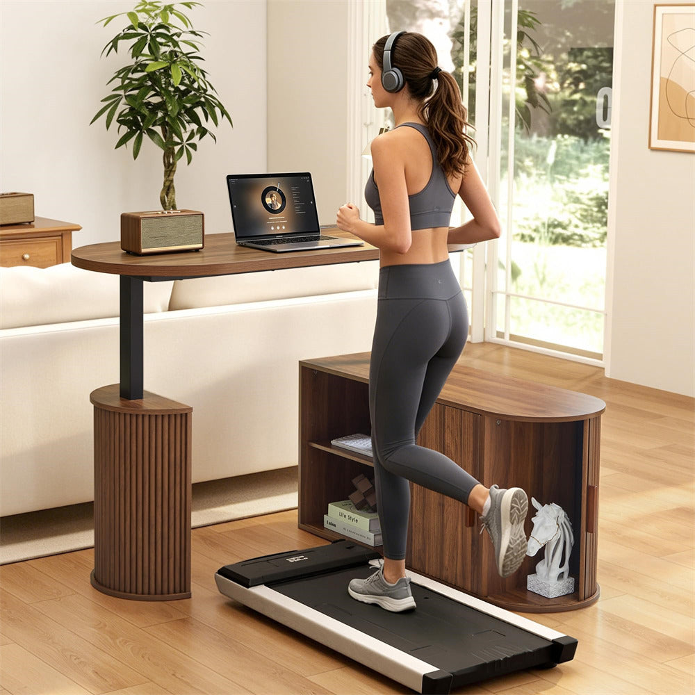 A woman in gray athletic wear runs on a black and silver treadmill in a bright, modern home office. She is positioned in front of a wooden standing desk with a laptop, a potted plant, and a curved wooden storage cabinet.