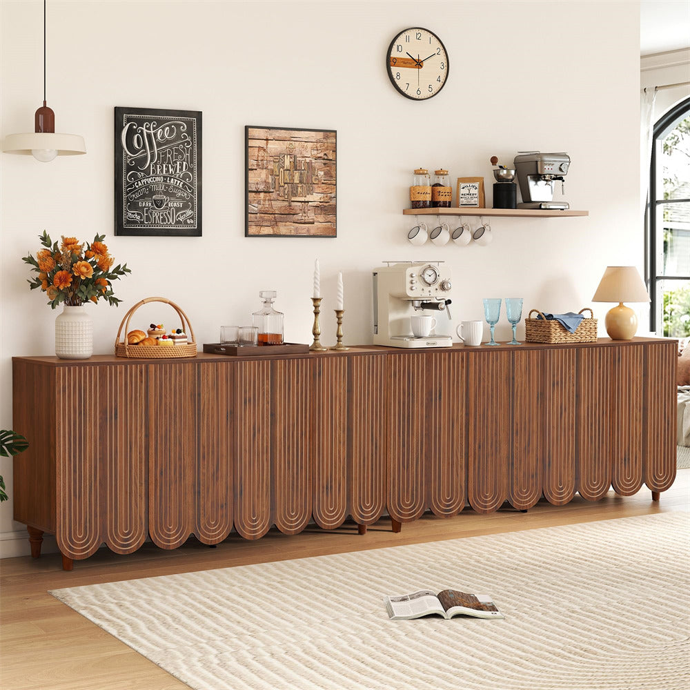 A long mid-century modern wooden sideboard or credenza with scalloped front panels, shown in a styled living room setting with wall art, a clock, shelves, coffee equipment, and a rug.