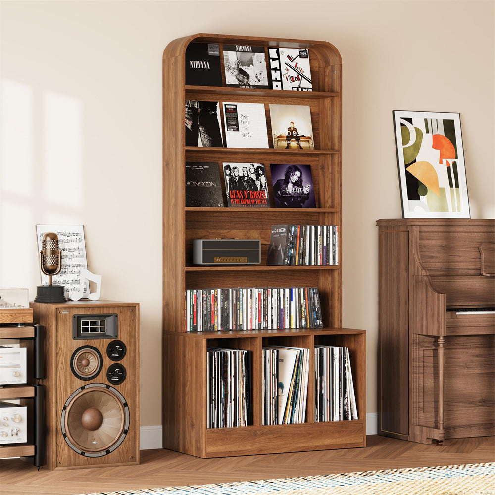 A tall wooden record and media shelf stands against a beige wall, holding vinyl records, CDs, and books. The room includes matching wood furniture, a speaker, a piano, and framed wall art.
