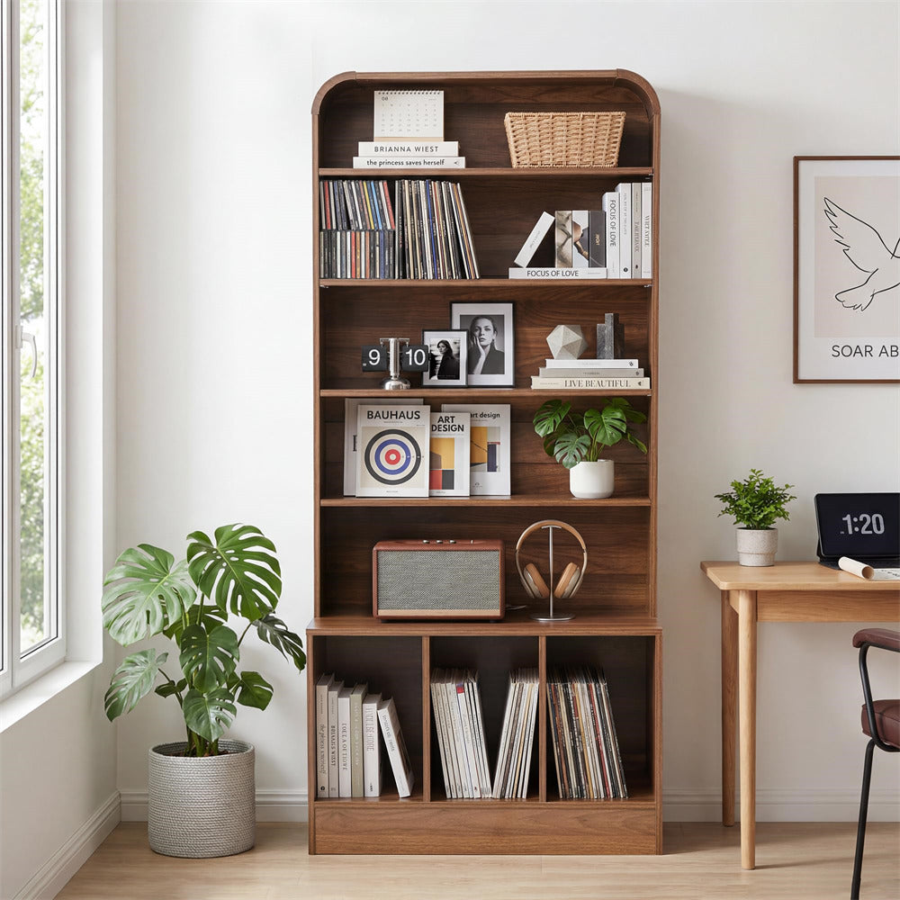 A tall wooden bookcase with five open shelves stands in a bright room beside a window, holding books, baskets, framed photos, plants, and small decor items. A small wooden desk with a plant and clock is visible on the right.