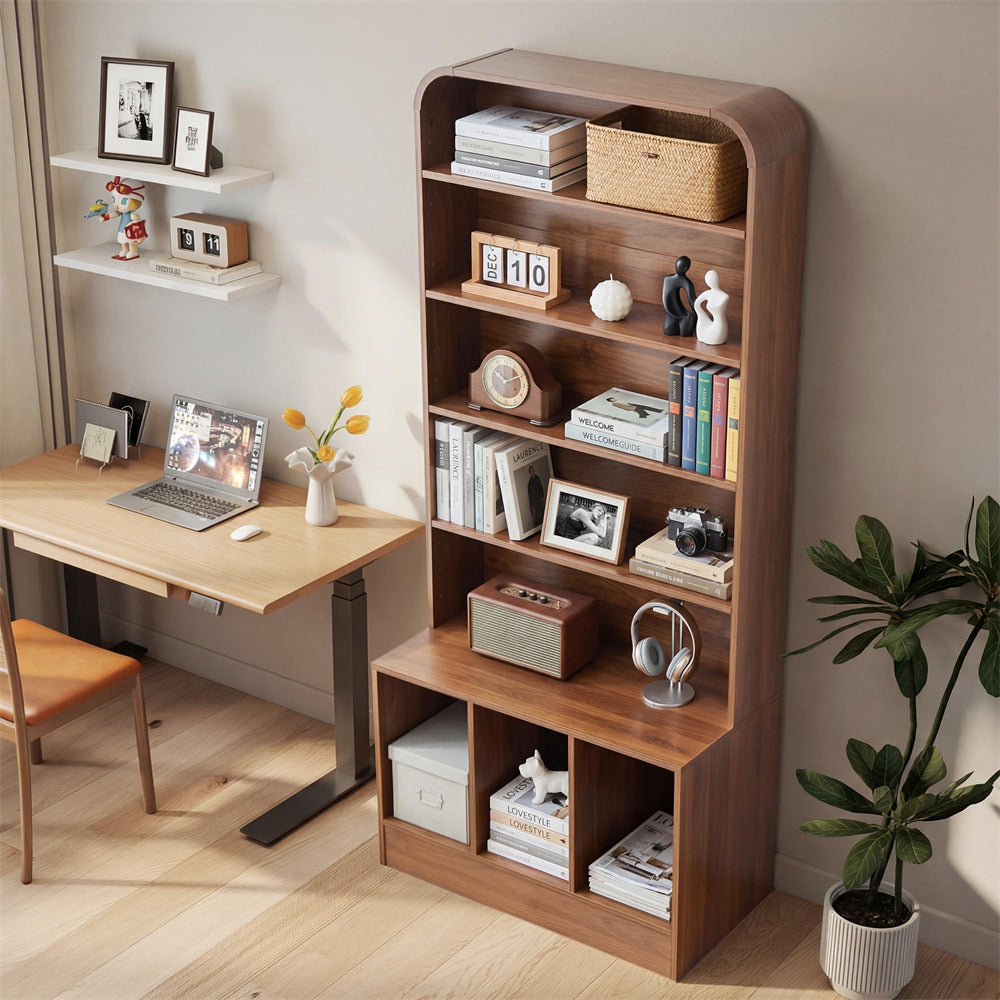 A tall wooden bookshelf with rounded top corners stands in a styled home office corner next to a desk. The bookshelf has five open shelves and two lower cubbies, filled with books, frames, a basket, a small speaker, headphones, and storage boxes.