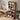 A tall wooden bookshelf with rounded top corners stands in a styled home office corner next to a desk. The bookshelf has five open shelves and two lower cubbies, filled with books, frames, a basket, a small speaker, headphones, and storage boxes.