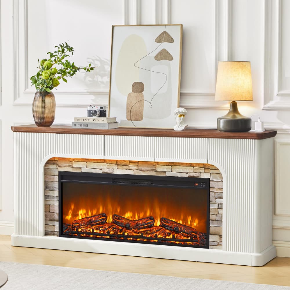 A white electric fireplace with a wood-look mantel and faux log flames, styled in a bright living room with a vase, framed art, books, and a table lamp.
