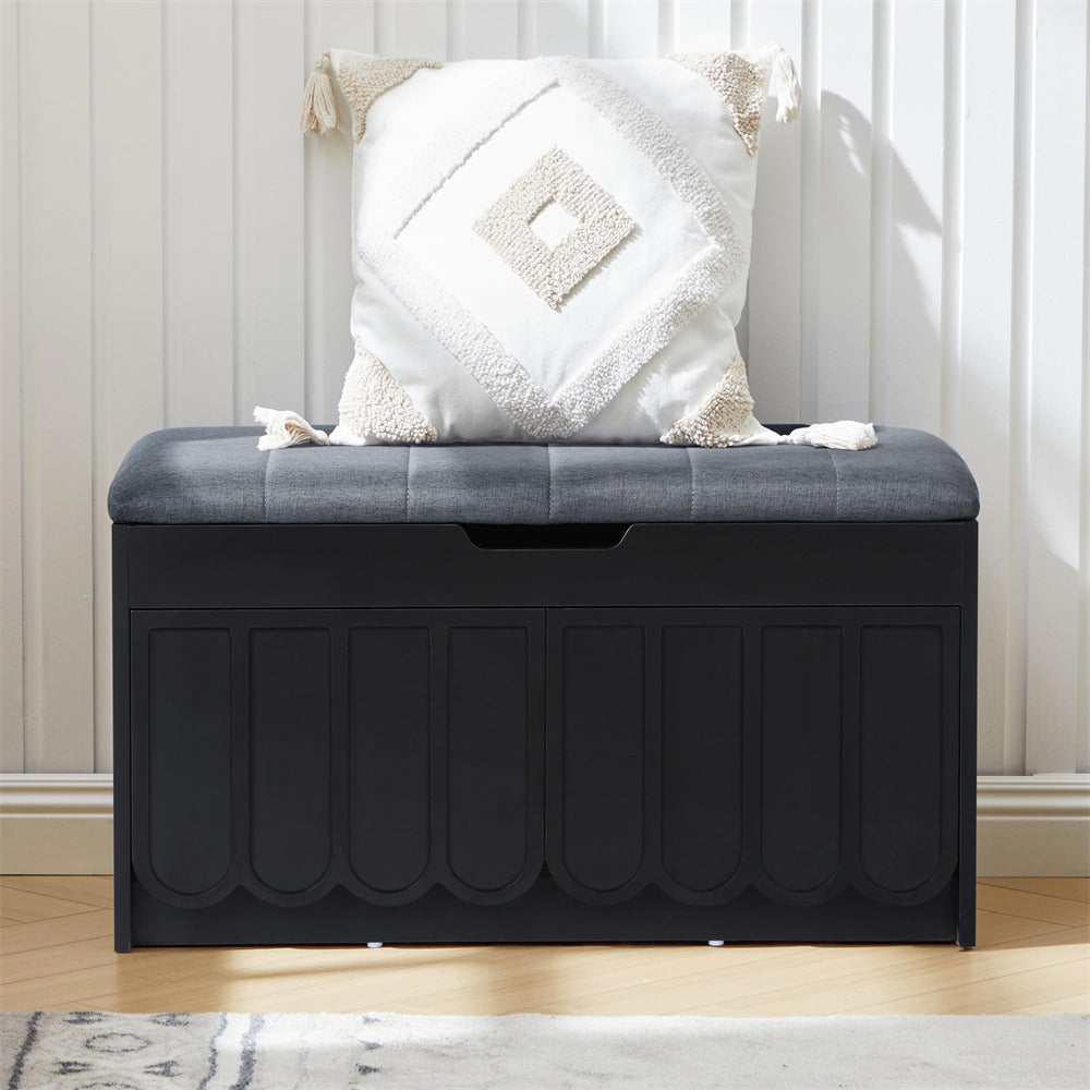Black storage bench with gray cushion and decorative pillow against a white paneled wall.