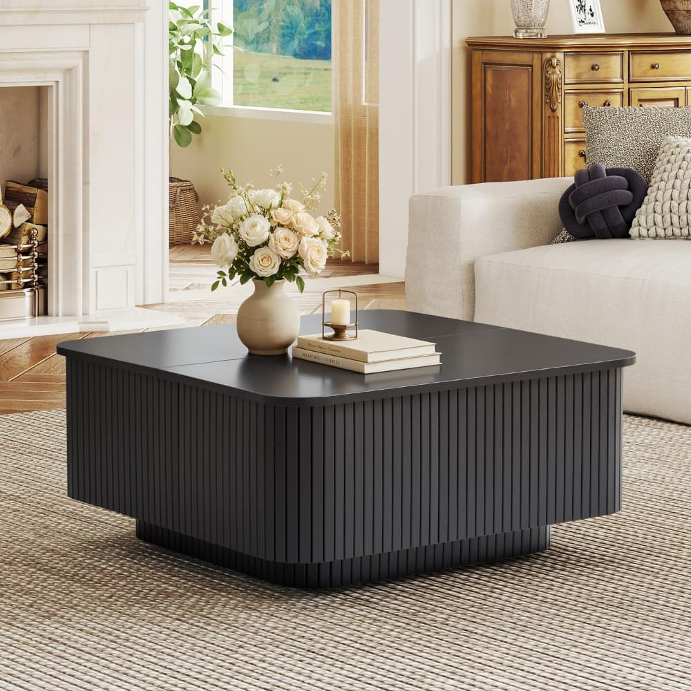 A modern black coffee table with a fluted front and rounded square shape, styled in a bright living room with a vase of flowers and books on top.