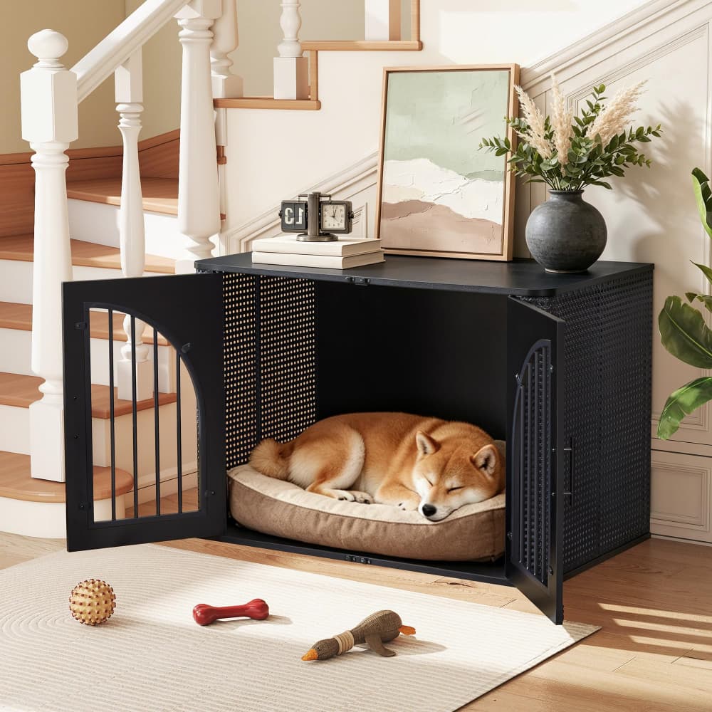 A black metal dog crate with an open door and mesh sides, shown in a staged home interior with a sleeping Shiba Inu dog inside on a cushioned bed. The crate is placed on a rug near toys, with a staircase and framed art in the background.