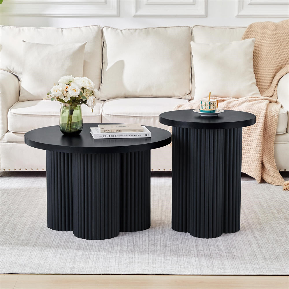 Two round black nesting coffee tables with fluted pedestal bases, styled in a bright living room with a cream sofa, white rug, and a peach throw blanket.