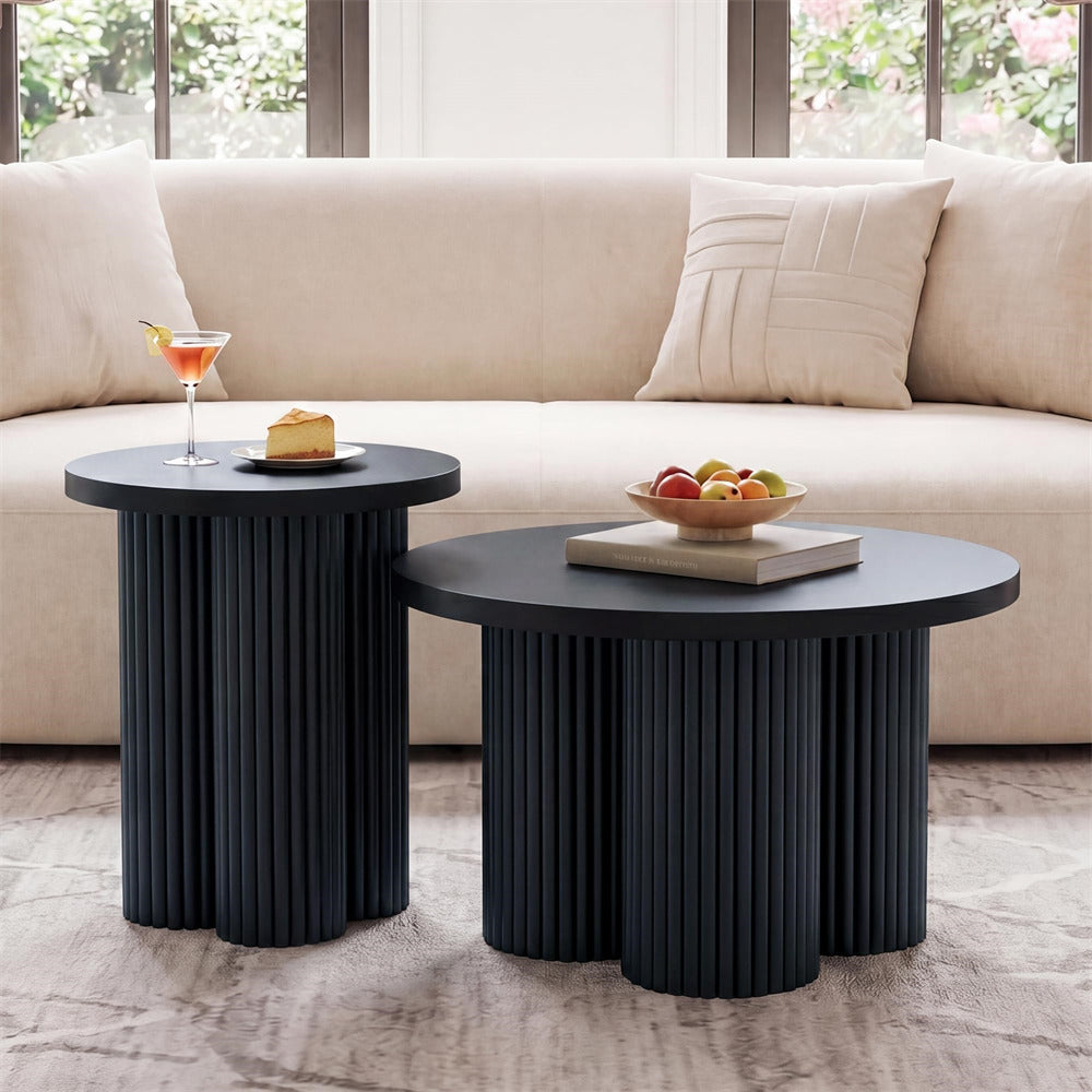 Two round nesting side tables with dark fluted pedestal bases, styled in a living room setting in front of a beige sofa. One table holds a martini glass with an orange cocktail and a slice of cake, while the other holds a bowl of fruit on a book.