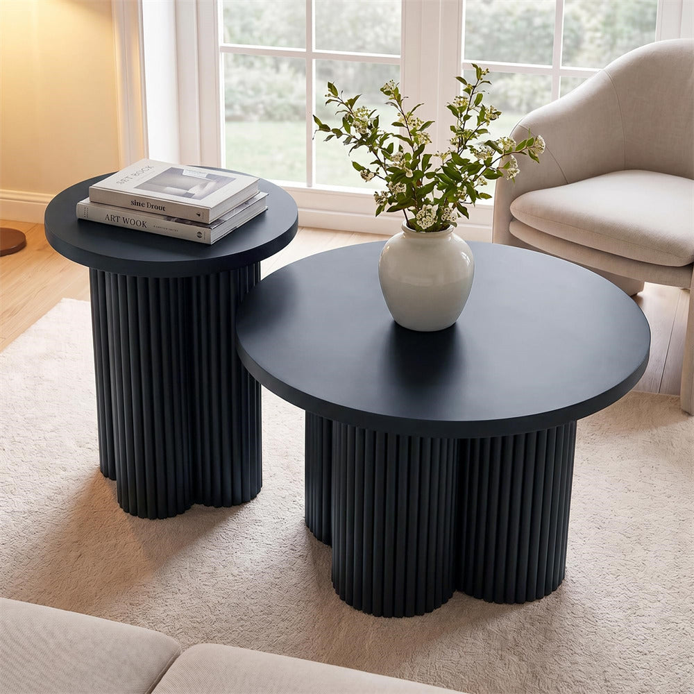 Two modern nesting coffee tables in a dark charcoal gray fluted base, styled in a bright living room with a white ceramic vase of greenery on the larger round table and books on the smaller side table.