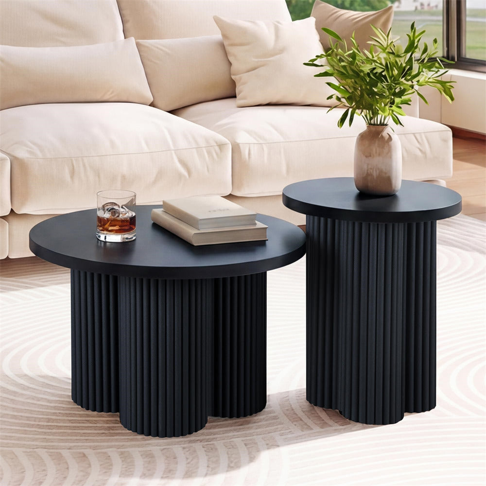 Two modern nesting side tables in matte black with fluted pedestal bases, styled in a bright living room with a cream sofa, a glass of whiskey, books, and a potted plant.