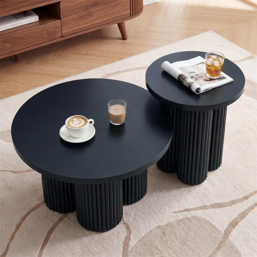 Two round nesting coffee tables in matte black with ribbed pedestal bases, styled on a light beige rug with a wooden cabinet in the background. One table holds a latte and a glass, and the other holds a rolled magazine and an iced drink.