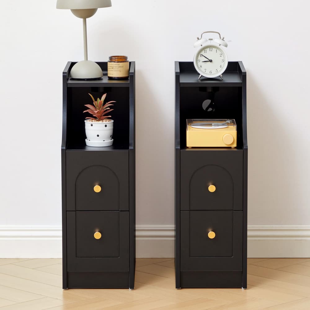 Two black narrow side tables with two drawers and gold knobs, styled with home decor items like a lamp, plant, and alarm clock, set against a plain white wall and light wood floor.