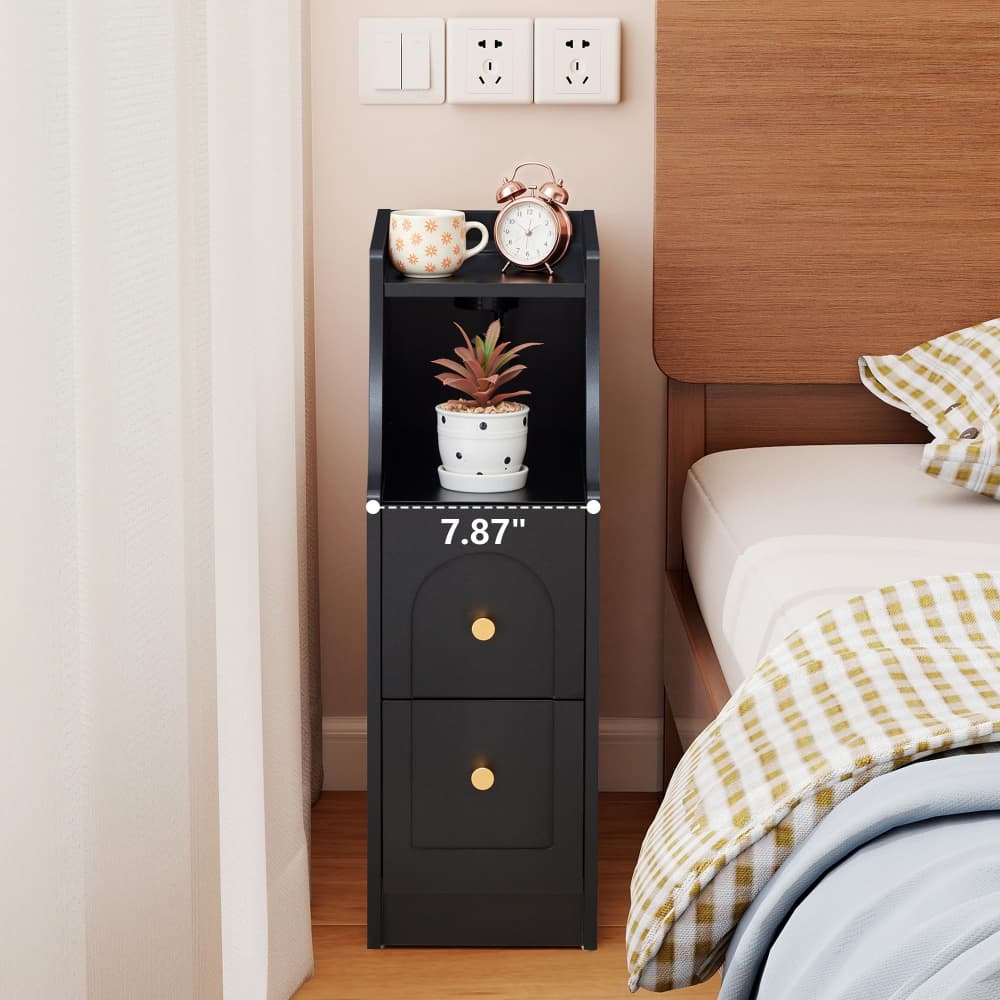 A black two-drawer nightstand with gold knobs stands beside a bed in a bedroom. The nightstand has open top shelves displaying a mug, a small alarm clock, and a potted succulent, with a dimension overlay reading 7.