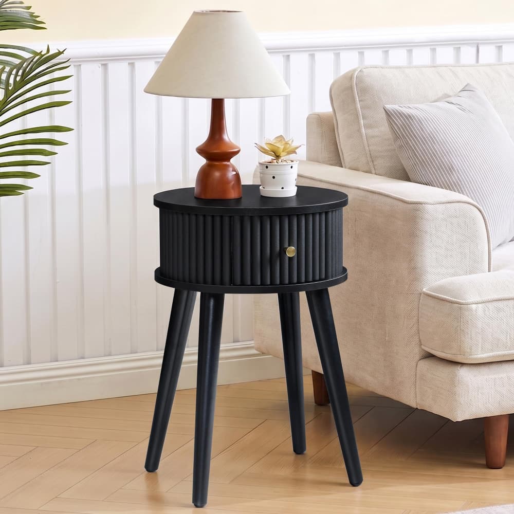 A black round side table with three tapered legs and a ribbed front, styled in a living room next to a beige armchair with a table lamp and small plant on top.