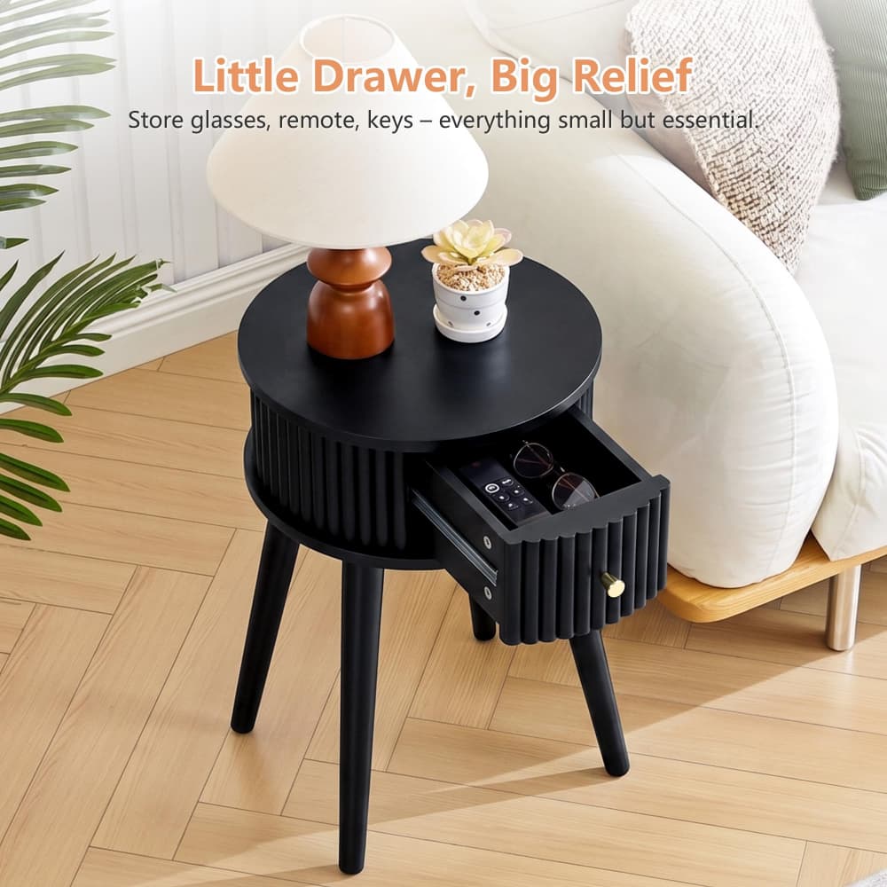 A black round side table with three legs and a ribbed drawer front, shown in a living room next to a sofa. One drawer is open, revealing stored items.