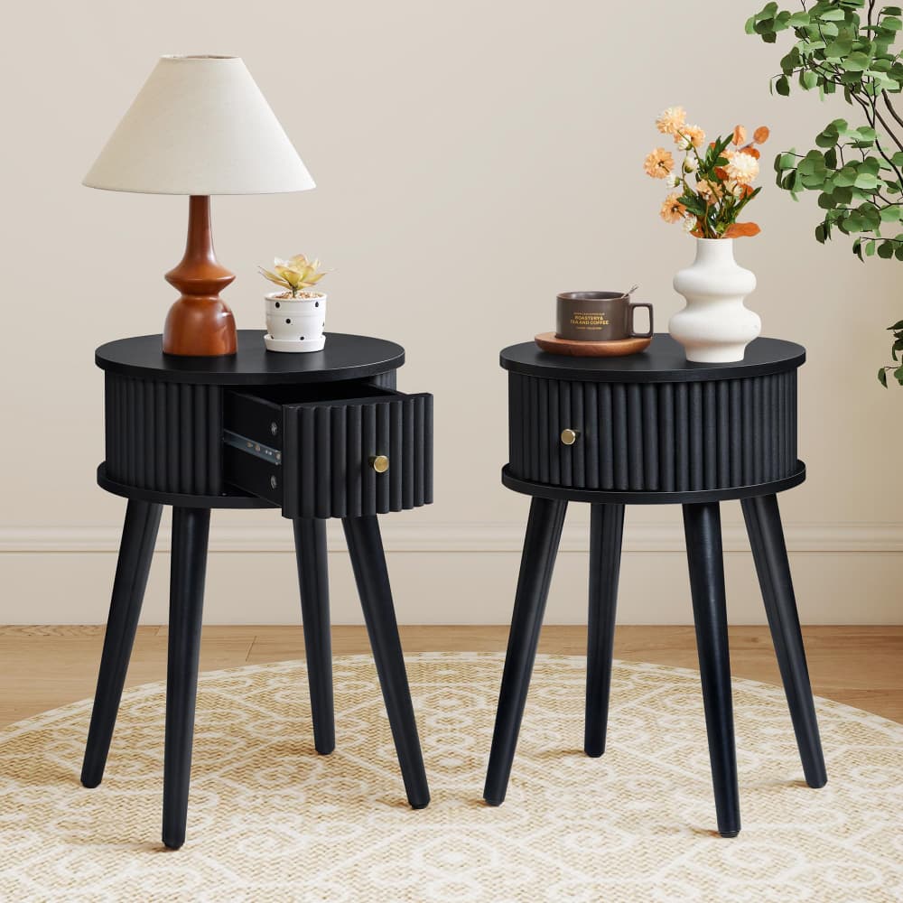 Two black ribbed side tables with tapered legs in a styled room setting. One table holds a table lamp and a small potted plant, while the other holds a white vase with flowers and a small cup and saucer.
