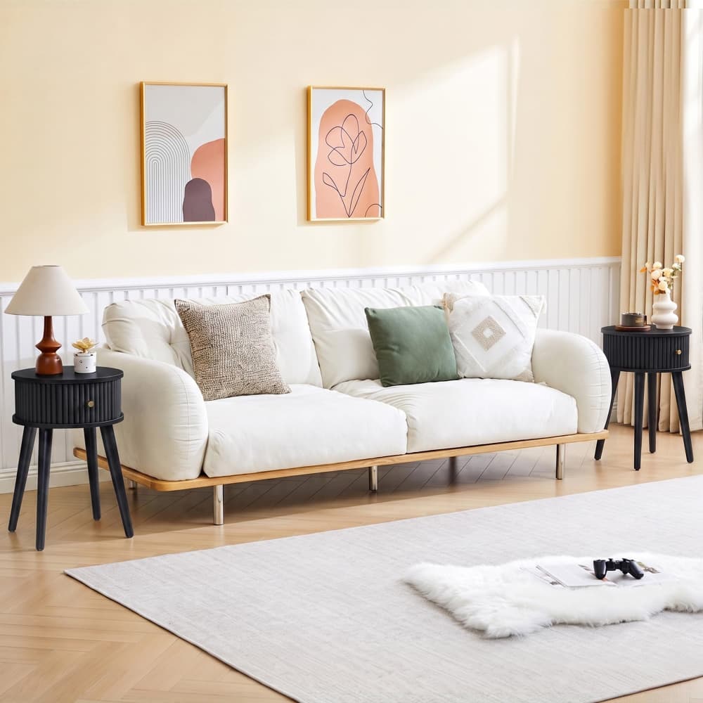 A cream-colored upholstered sofa with a light wood base and legs, styled in a warm, modern living room with two side tables, a lamp, and two framed abstract art prints on the wall.