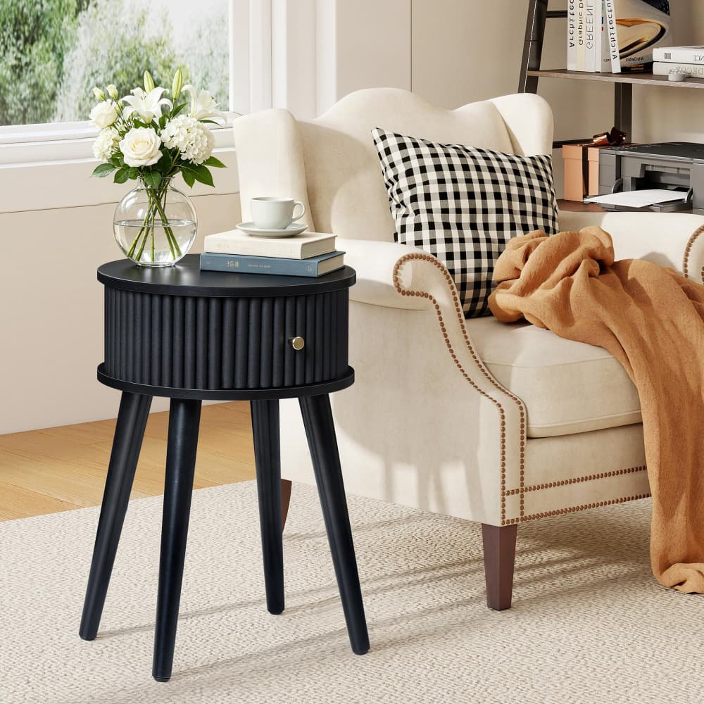 A black round side table with three legs and a ribbed front, styled in a bright living room next to a cream armchair with a black-and-white check pillow and a tan throw blanket.