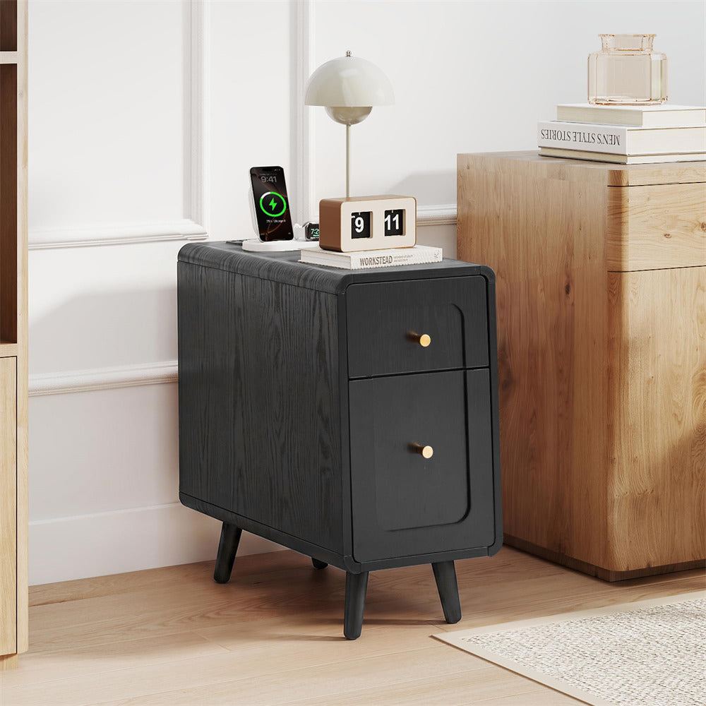 A black wood-grain nightstand with two drawers and gold knobs, styled in a bright room next to a wooden cabinet.