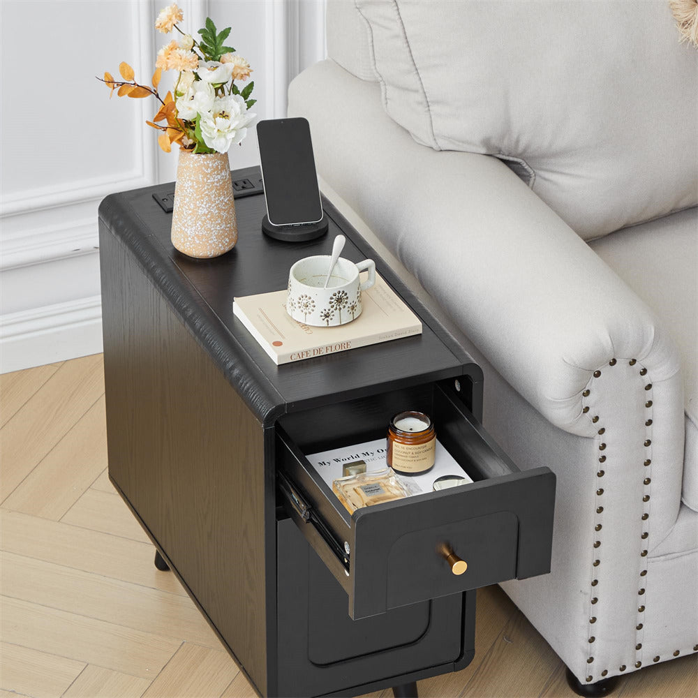 A black side table with an open drawer and a top tray, styled next to a light gray sofa. The table is decorated with a vase of flowers, a smartphone, a book, a cup and saucer, and a small jar, set on a light wood floor.
