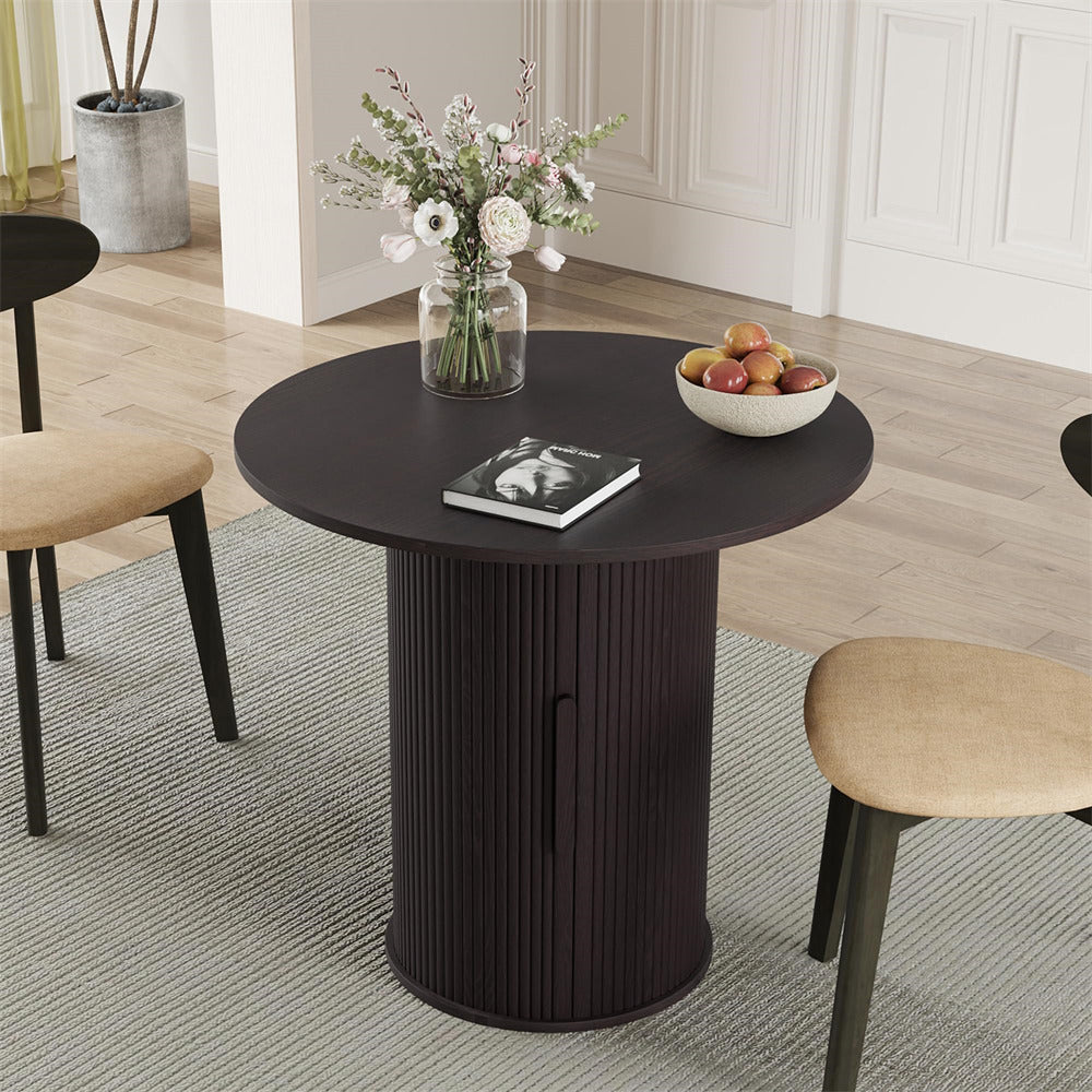 Round black table with a vase of flowers, book, and bowl of fruit in a room with chairs and a rug.