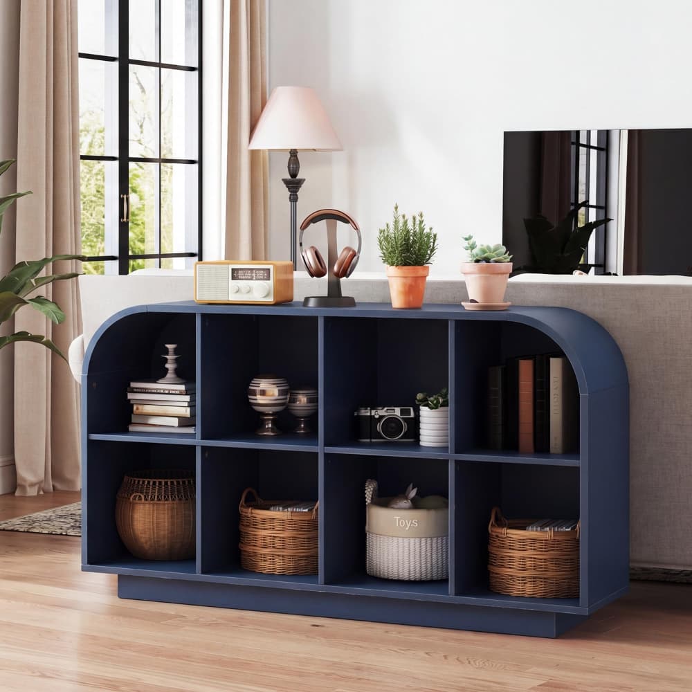 A navy blue arched media console with nine open cubbies, styled in a bright living room with books, baskets, a lamp, and potted plants.