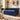 A blue velvet Chesterfield loveseat sofa with tufted backrest, rolled arms, nailhead trim, and dark turned legs, styled in a bright living room with pillows, a throw blanket, rug, plants, and wall decor.