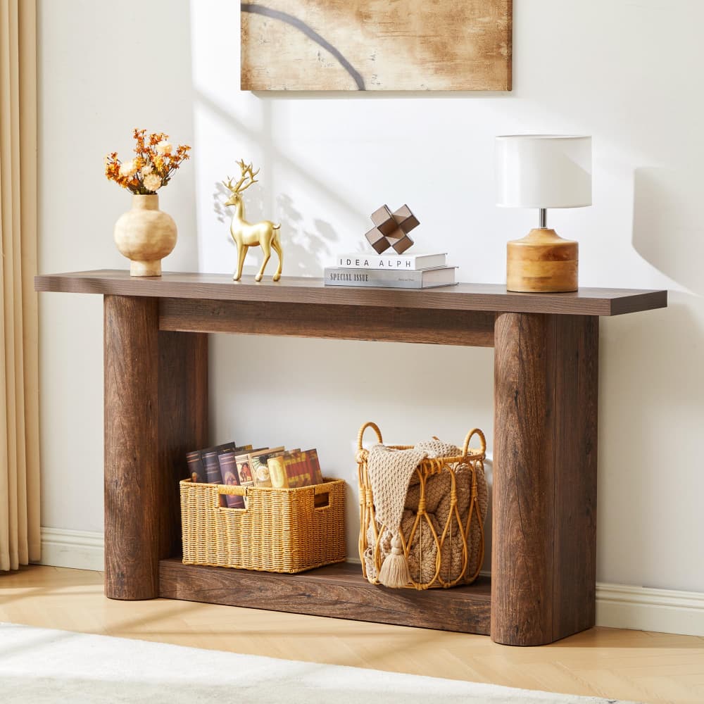A rustic dark brown wooden console table styled in a bright, modern room with a white wall, wooden floor, and decorative items including a lamp, vase, books, and baskets.