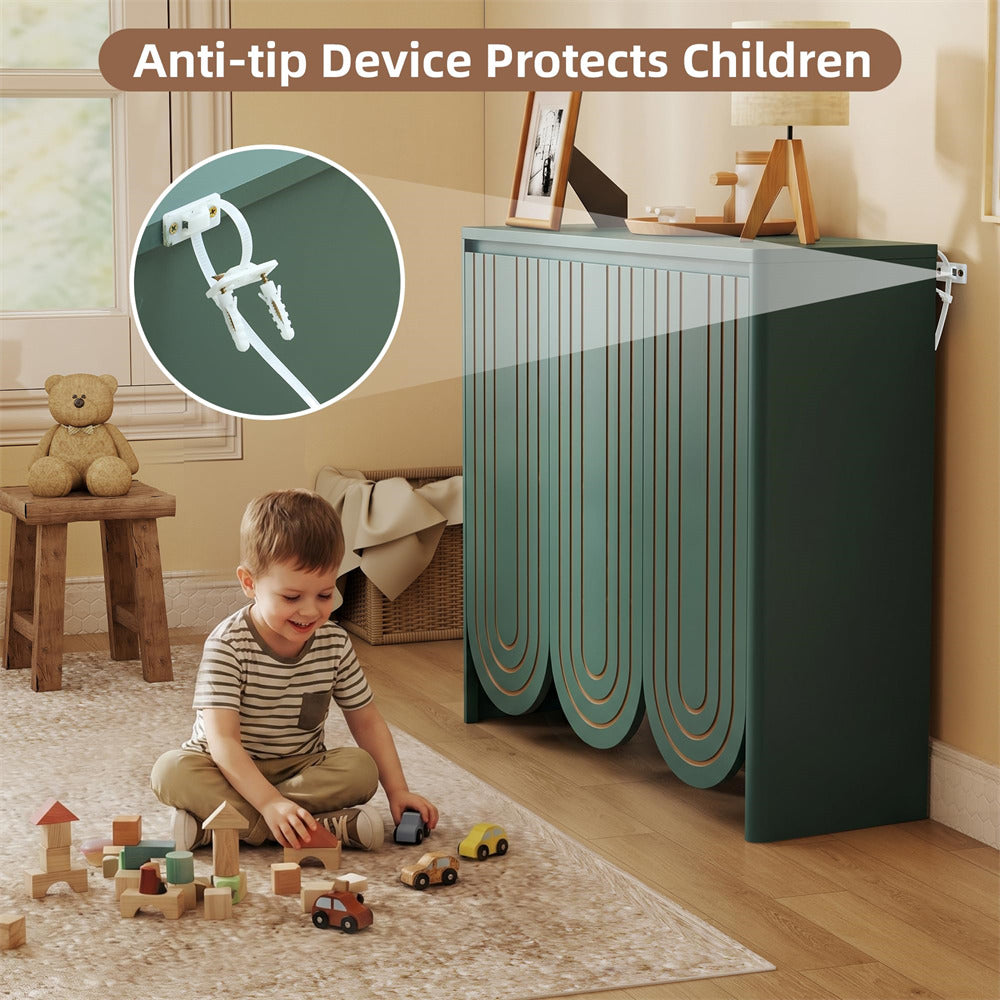 A children's storage cabinet in a warm beige room, with a young boy sitting on the floor playing with toys. The cabinet has a scalloped front design in muted teal and dark green, with a white anti-tip device visible on the top right corner.