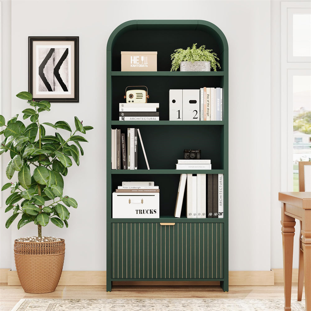 A tall dark green bookcase with arched top, four open shelves, and a slatted lower cabinet door, styled with books, binders, a small plant, and decorative items in a bright room.