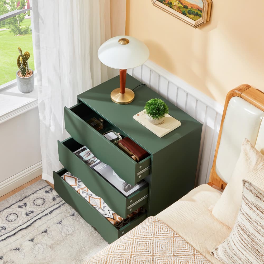 A dark green three-drawer nightstand in a bedroom setting, styled with a white table lamp, a small potted plant, and open drawers showing storage space.