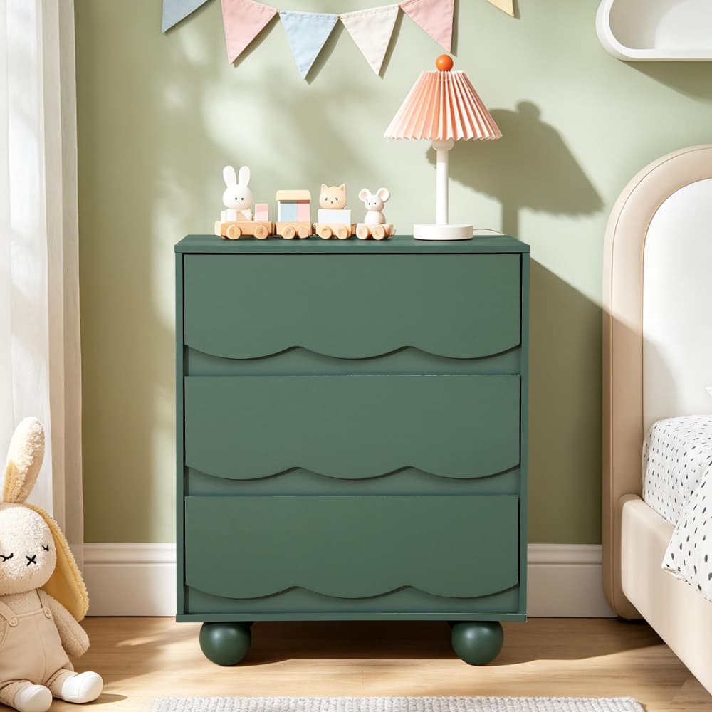 A dark green three-drawer dresser with scalloped drawer fronts and round feet, styled in a child's bedroom next to a bed with a pink pleated lamp and small toys on top.