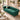 A plush emerald green velvet Chesterfield-style sofa with button tufting, nailhead trim, and dark wooden legs, styled in a bright, elegant living room with beige walls, a marble coffee table, and neutral throw pillows.