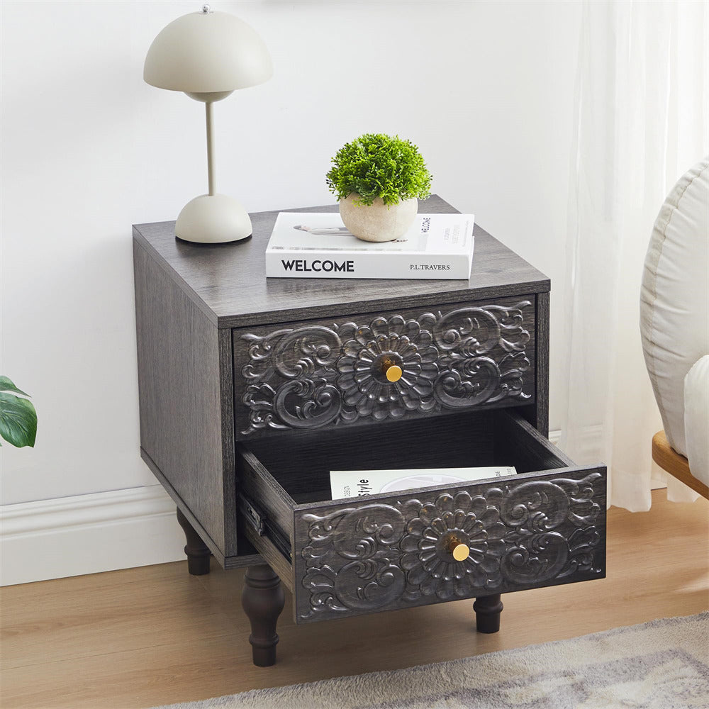 Grey Decorative nightstand with embossed design, open drawer, books, plant, and lamp on a wooden floor.