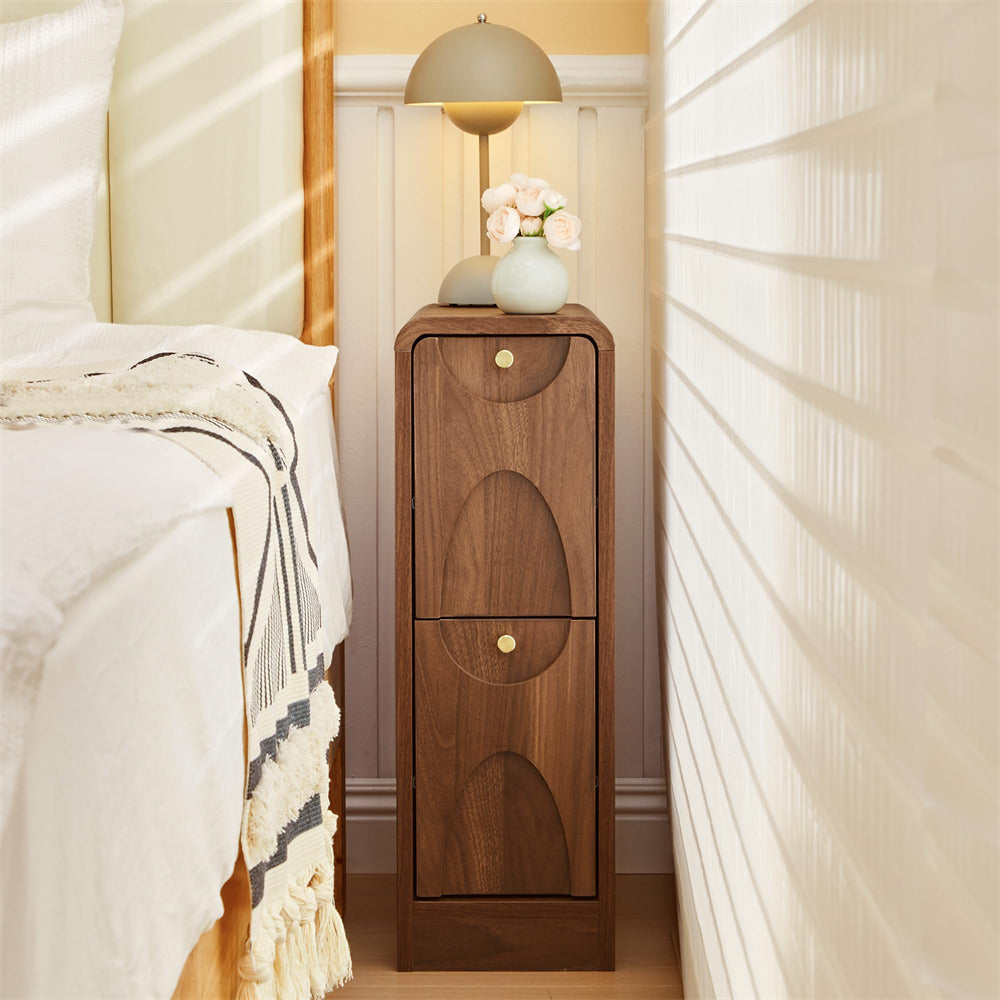 A tall wooden nightstand with two arched drawer fronts and small round gold knobs, styled in a bedroom next to a bed with white bedding. On top of the nightstand are two dome-shaped lamps and a small vase with flowers.