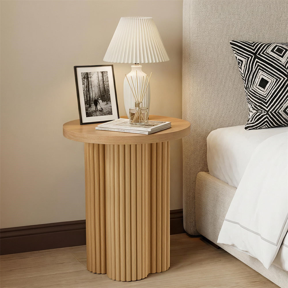 Round wooden side table with lamp and decor items next to a bed with a patterned pillow.