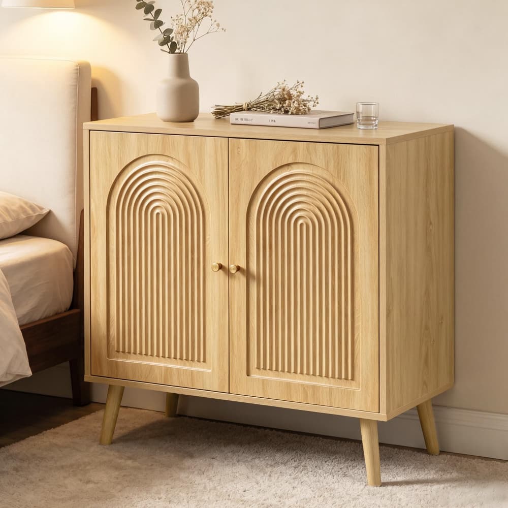 A light natural wood nightstand with two arched door panels and small angled legs, styled in a bedroom next to a bed with a vase, book, and glass on top.