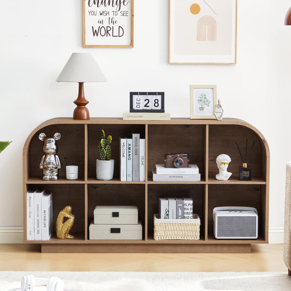 A wooden arched bookshelf with open cubbies styled with books, a small lamp, decorative figurines, a potted cactus, and storage boxes in a bright, modern room.