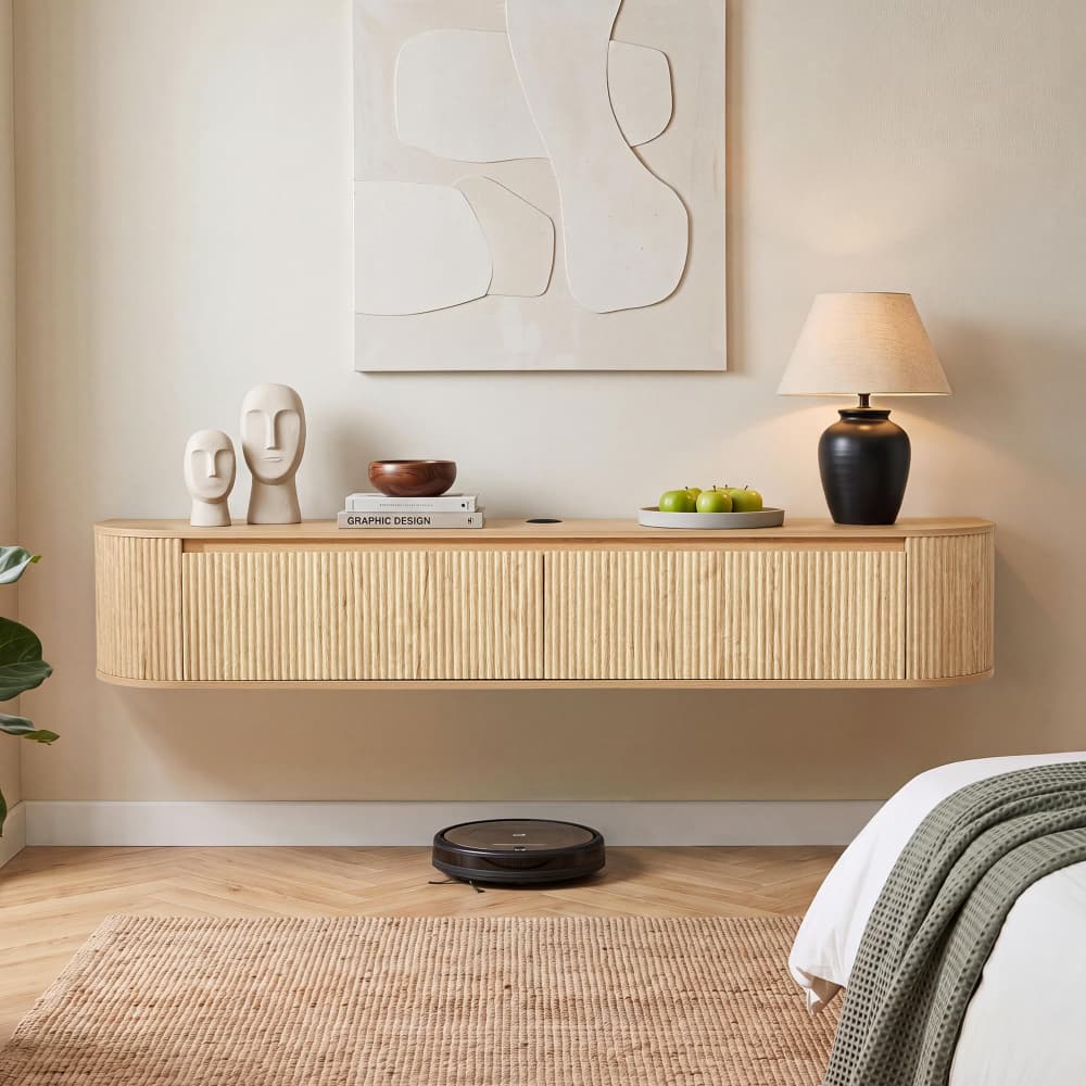 A modern floating wooden console shelf mounted on a beige wall in a styled bedroom or living space, holding decorative objects and a lit table lamp. A bed with a green knit throw is partially visible in the foreground, along with a woven rug and a robotic vacuum on the floor.