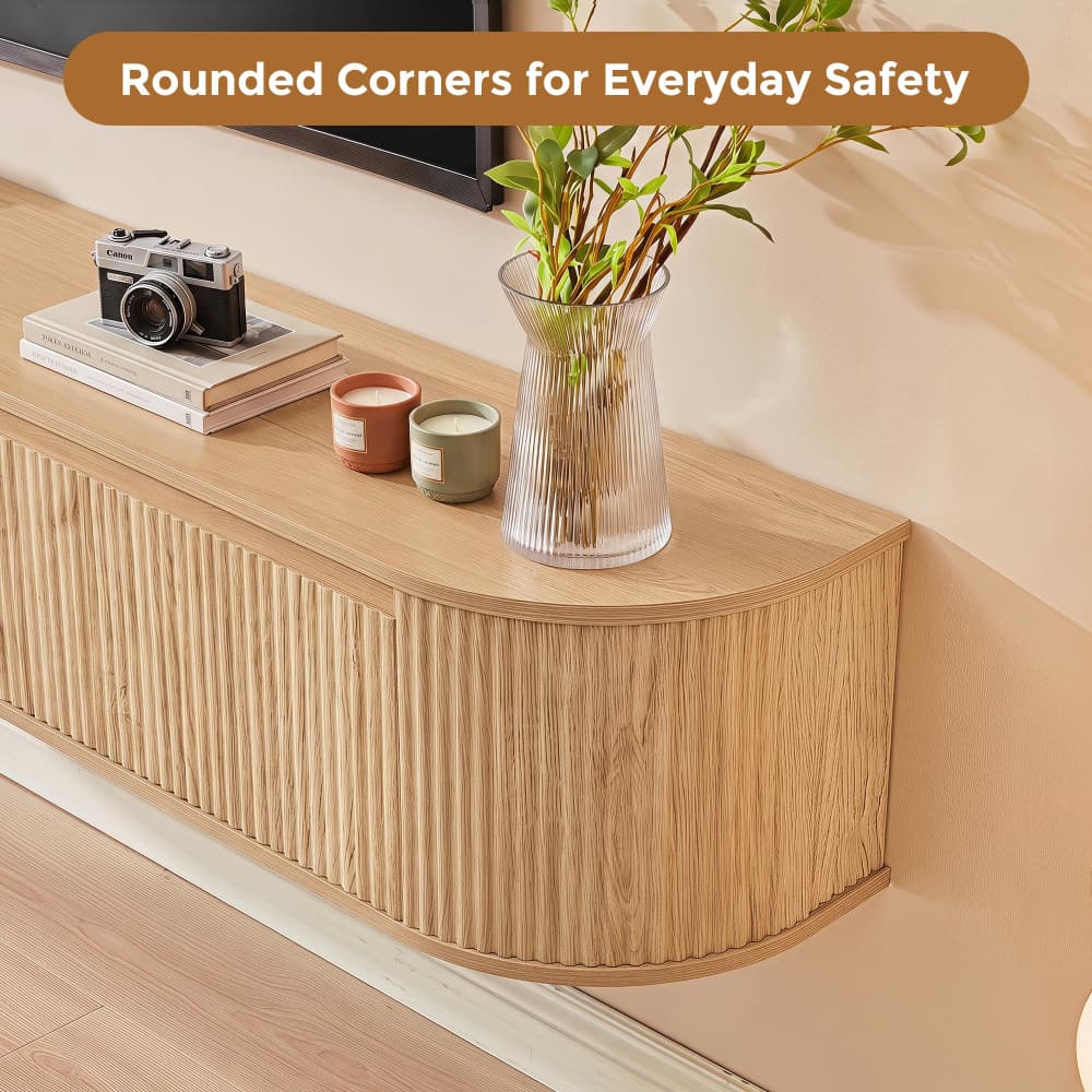 A light wood floating TV console with a fluted front panel and rounded corners, styled with a glass vase of greenery, a vintage camera, and candles against a beige wall.