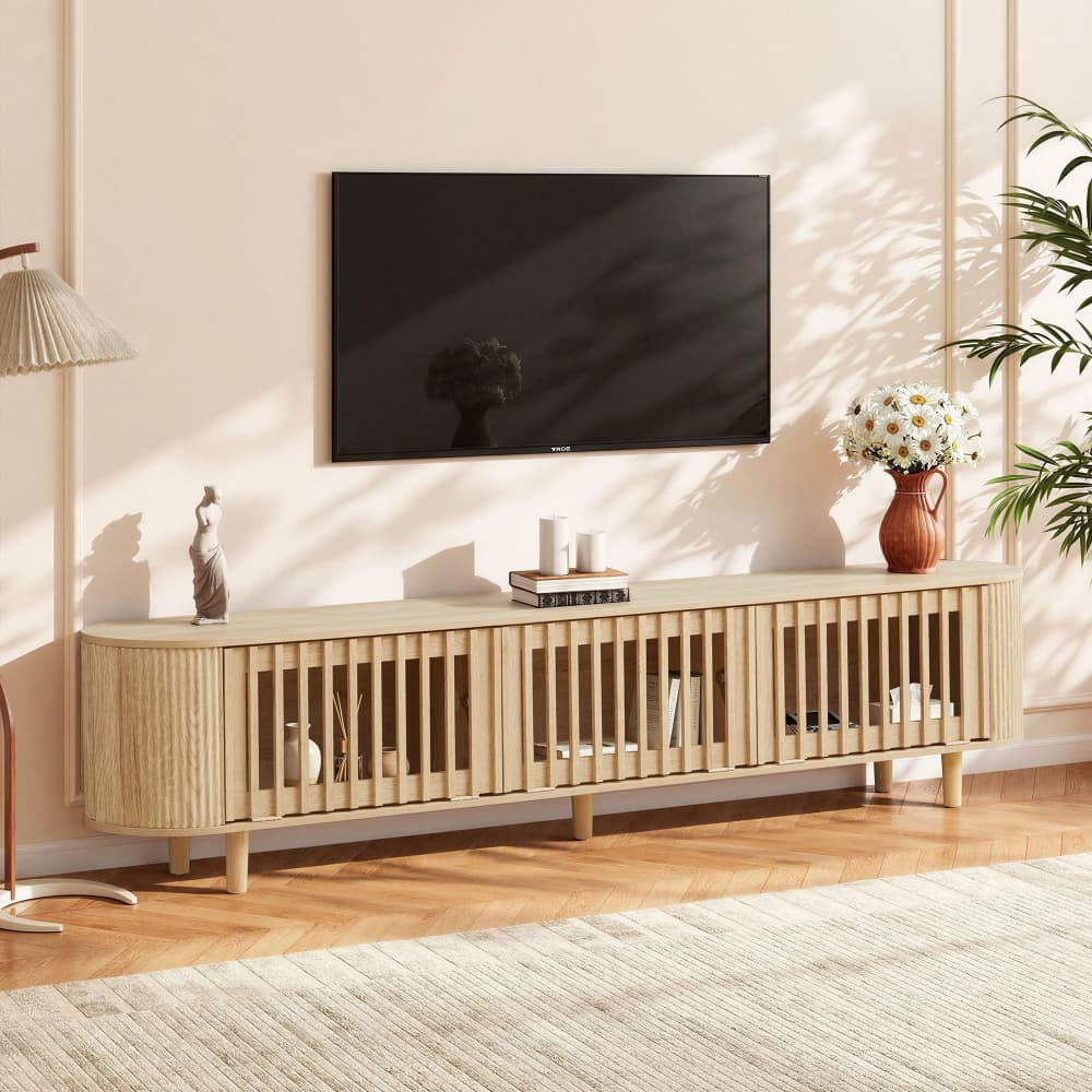 A modern light wood TV console in a styled living room setting, featuring a black flat-screen TV mounted on the wall above it, with decorative items and a vase of flowers on top.